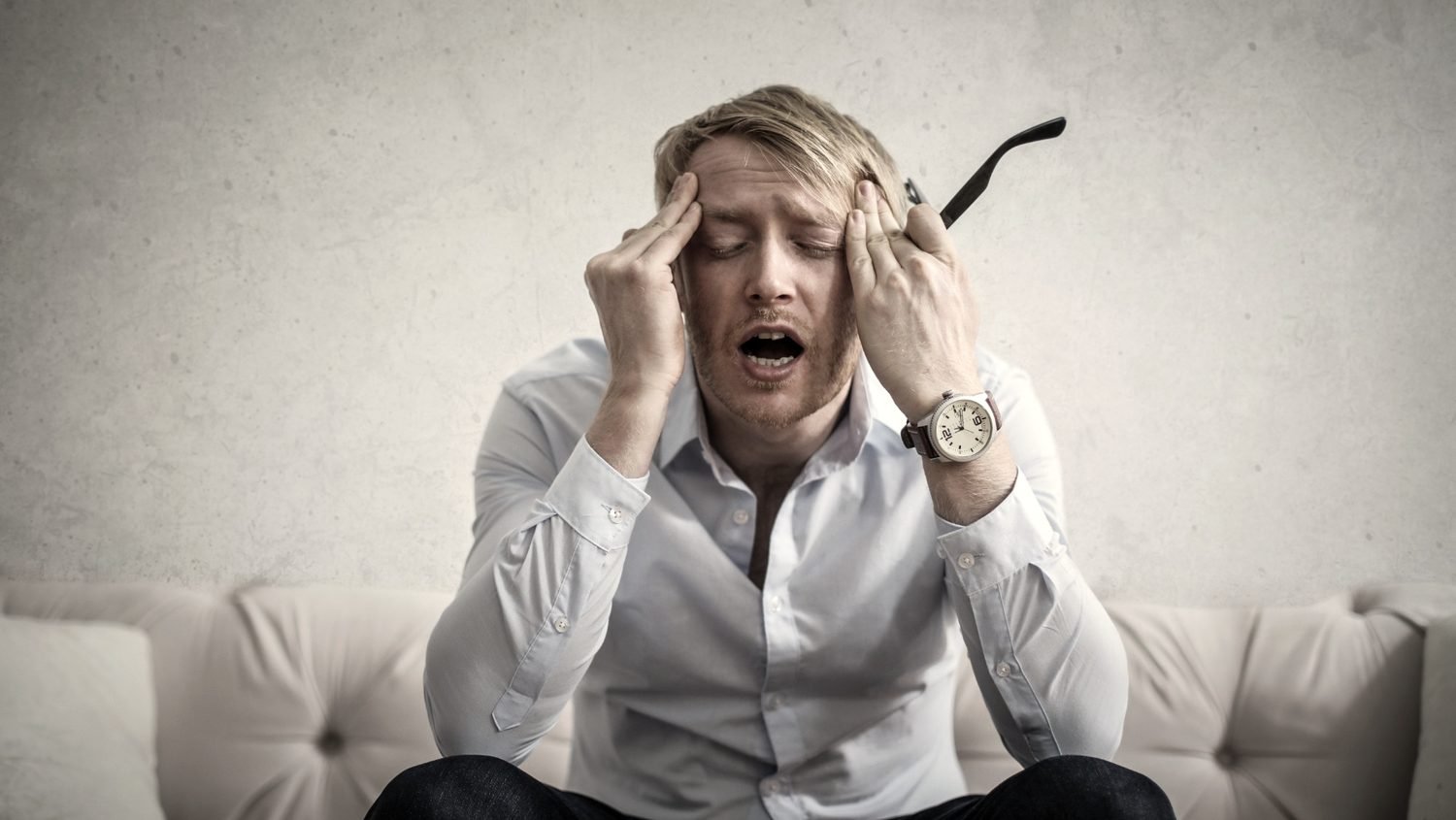 Un homme assis sur un canapé, les mains aux tempes, épuisé par une douleur qui dure depuis des mois